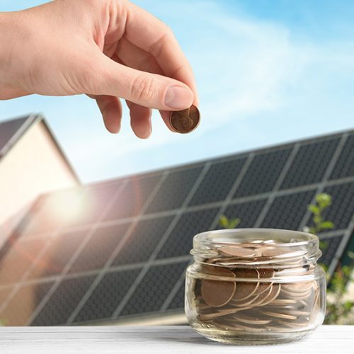 Woman putting coin into jar against house with installed solar panels on roof, closeup. Economic benefits of renewable energy